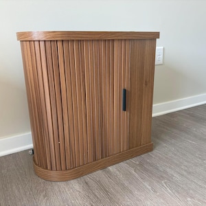 Puede incluir: Un mueble de madera con un diseño de listones verticales y una base curva. El mueble es de color marrón cálido y tiene un tirador negro. El mueble está colocado contra una pared blanca y un suelo gris claro.