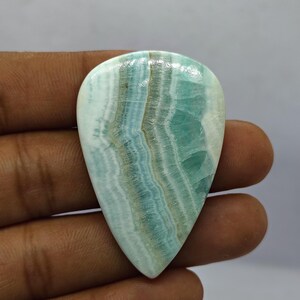 May include: A polished, teardrop-shaped gemstone with a mix of light blue, white, and brown bands. The stone has a glossy surface and a smooth texture. The gemstone is held in a hand, showcasing its natural beauty and unique pattern.