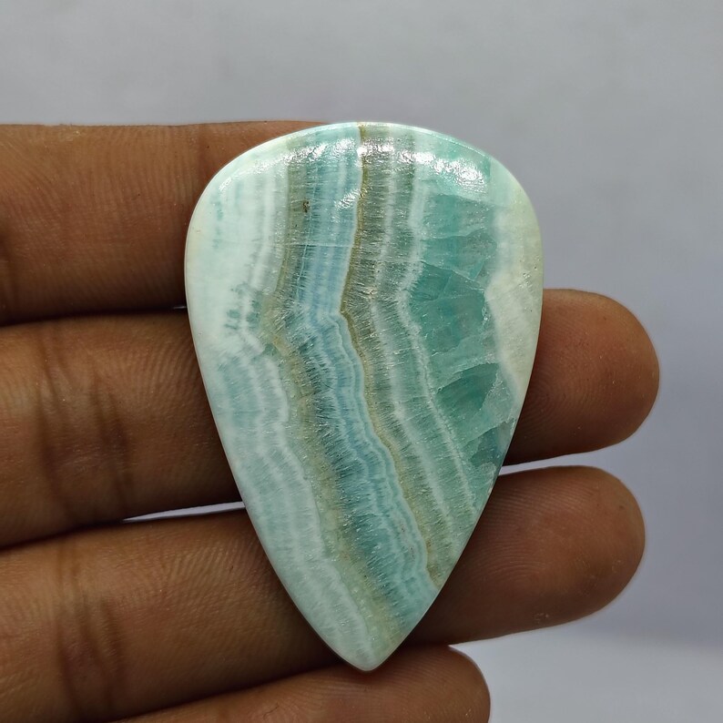 May include: A polished, teardrop-shaped gemstone with a mix of light blue, white, and brown bands. The stone has a glossy surface and a smooth texture. The gemstone is held in a hand, showcasing its natural beauty and unique pattern.