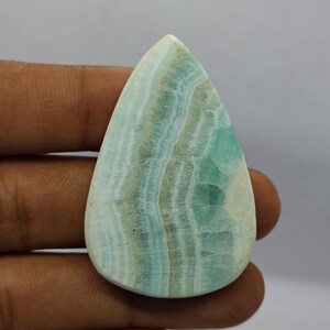 May include: A teardrop-shaped gemstone with a polished surface. The stone features bands of light blue, teal, and white, creating a striped pattern. The gemstone is held in a hand, showcasing its size and shape.