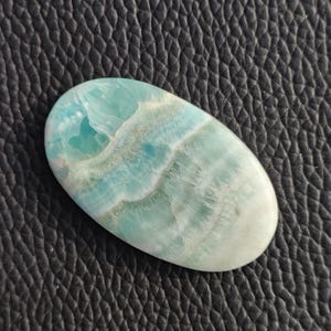 May include: An oval-shaped gemstone with a light blue and white pattern. The stone has a polished surface and a smooth texture. The color transitions from a pale blue to a creamy white. The gemstone is set against a black, textured background.