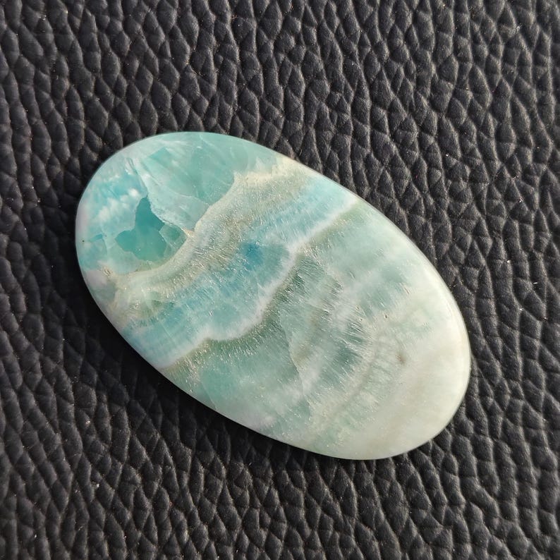 May include: An oval-shaped gemstone with a light blue and white pattern. The stone has a polished surface and a smooth texture. The color transitions from a pale blue to a creamy white. The gemstone is set against a black, textured background.