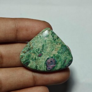 Puede incluir: Una piedra preciosa pulida de forma triangular que presenta una superficie verde moteada con distintas inclusiones de color rosa oscuro y púrpura. La piedra tiene una textura suave y ligeramente reflectante con un patrón natural de vetas minerales.