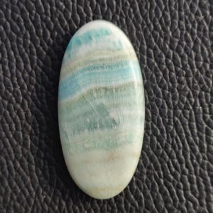 May include: An oval-shaped gemstone with a polished surface. The stone features layers of light blue, teal, and white hues, creating a striped pattern. The gemstone is set against a dark, textured background.