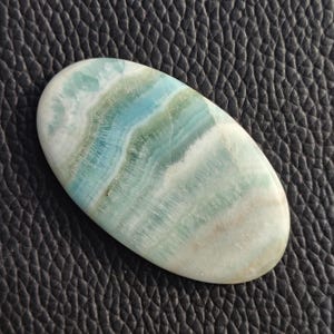 May include: Oval-shaped polished gemstone with a swirling pattern of blue, green, and white hues. The stone has a smooth, glossy surface and appears to be a natural mineral. The gemstone is set against a dark, textured background.