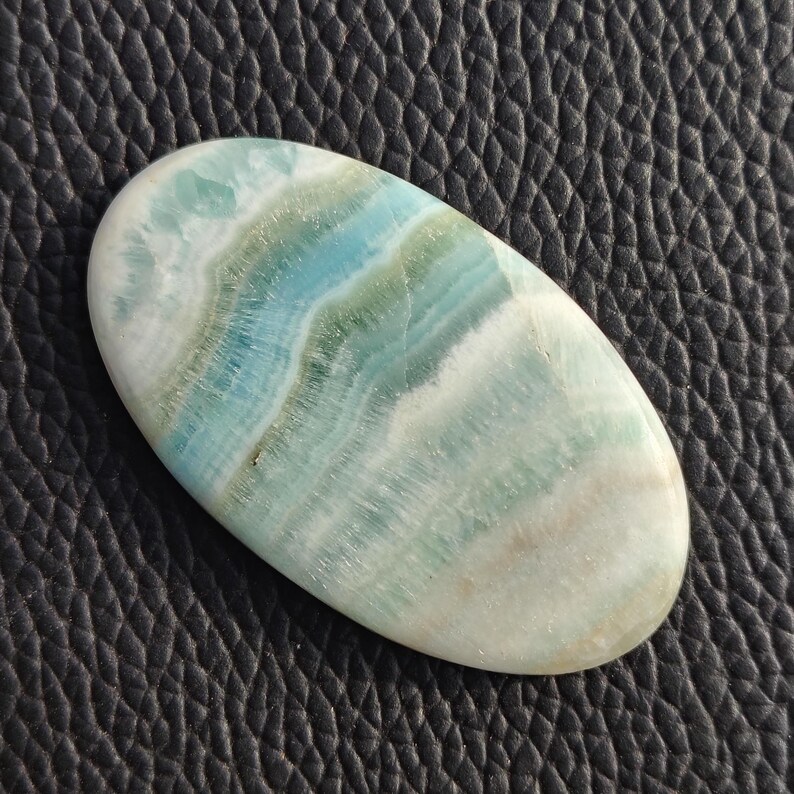 May include: Oval-shaped polished gemstone with a swirling pattern of blue, green, and white hues. The stone has a smooth, glossy surface and appears to be a natural mineral. The gemstone is set against a dark, textured background.