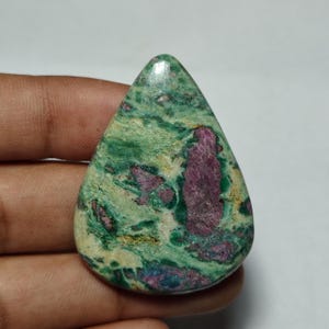 Puede incluir: Una piedra preciosa pulida en forma de lágrima que presenta un patrón jaspeado de colores verde vibrante y crema con distintas manchas irregulares de inclusiones de color magenta intenso y púrpura en toda la superficie lisa y brillante.