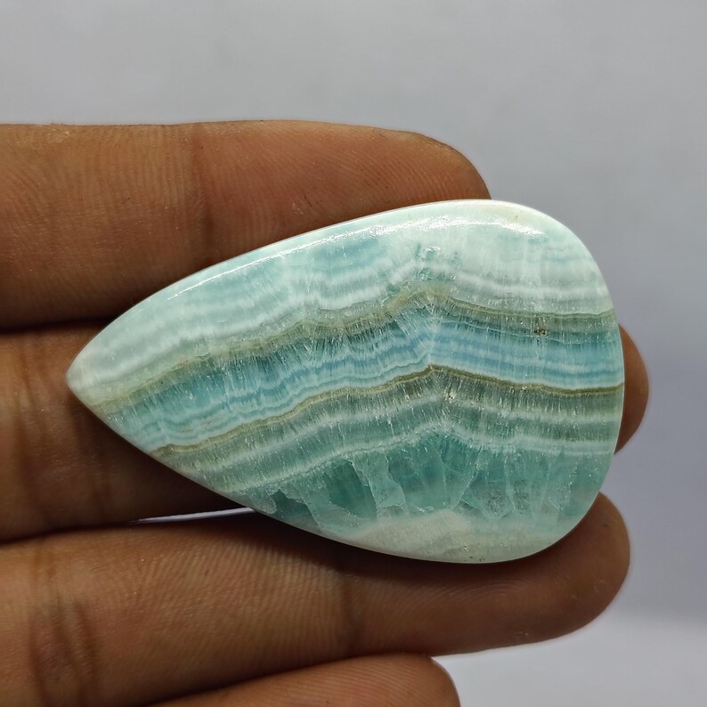 May include: A polished, teardrop-shaped gemstone with a layered pattern of light blue, white, and brown bands. The stone has a smooth, glossy surface and is held in a hand. The gemstone is likely used for jewelry or decorative purposes.