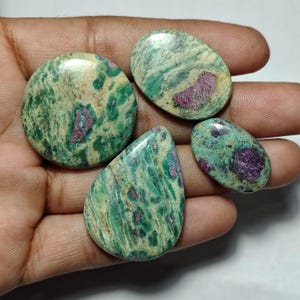 May include: Four polished cabochon gemstones featuring a mottled green and cream matrix with distinct, embedded magenta-coloured inclusions. The stones vary in shape, including circular, oval, and teardrop forms, displaying a smooth, glossy surface texture.