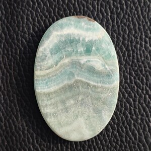 May include: Oval-shaped gemstone with a layered pattern of light blue, white, and gray hues. The polished surface of the stone displays a natural, flowing design. The gemstone is set against a dark, textured background.