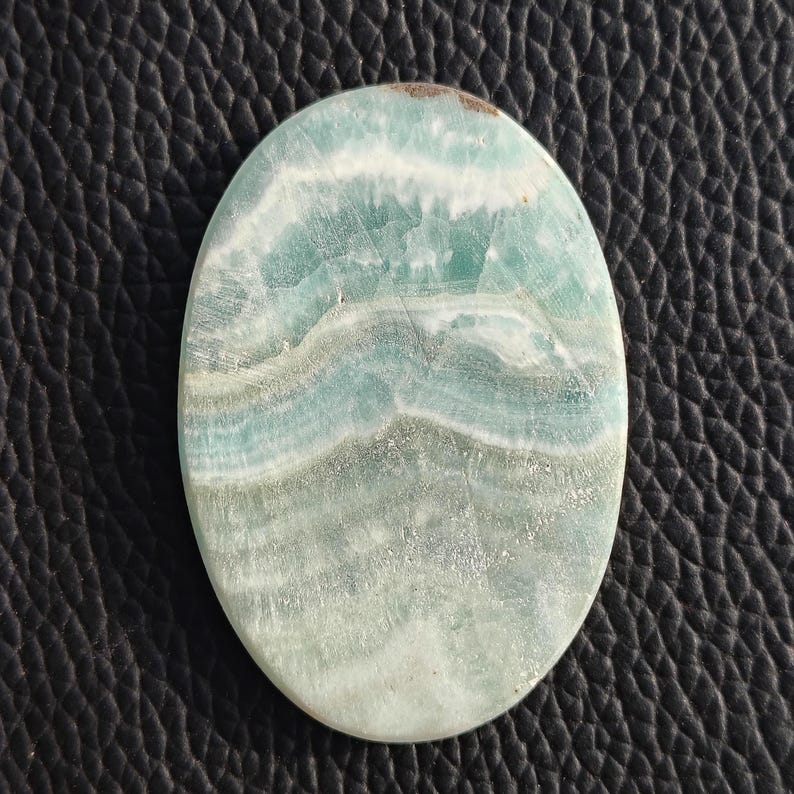 May include: Oval-shaped gemstone with a layered pattern of light blue, white, and gray hues. The polished surface of the stone displays a natural, flowing design. The gemstone is set against a dark, textured background.