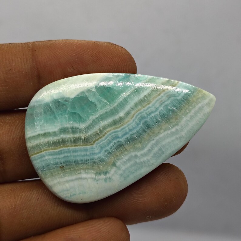 May include: A polished, teardrop-shaped gemstone with a captivating pattern of layered blue-green and white bands. The stone's surface is smooth, showcasing a natural, organic design. The gemstone is held in a hand.
