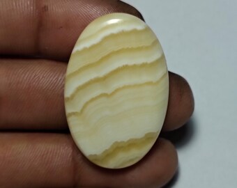 Puede incluir: Gema ovalada con una base blanca cremosa y bandas superpuestas de amarillo claro y marrón claro. La piedra pulida se sostiene entre los dedos, mostrando su superficie lisa y brillante. La gema mide aproximadamente 3,8 cm de largo.