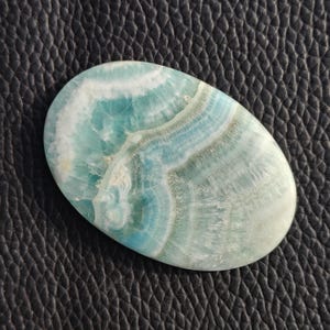 May include: Oval-shaped gemstone with a swirling pattern of blue, white, and gray hues. The polished stone has a smooth surface and a marbled appearance, showcasing natural variations in color and texture. The gemstone is set against a dark, textured background.