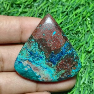 May include: A triangular-shaped gemstone with a polished surface. The stone features a blend of colors, including deep reds, blues, and turquoise, creating a marbled effect. The gemstone is held in a hand against a backdrop of green grass.