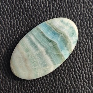 May include: An oval-shaped gemstone with a polished surface. The stone features a blend of light blue, white, and pale green hues, with a layered, wave-like pattern. The gemstone is set against a black textured background.
