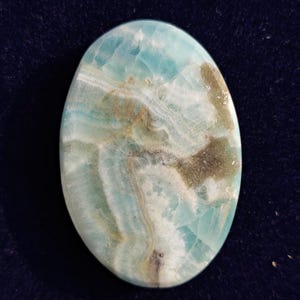 Op de afbeelding: Ovale edelsteen met een mix van lichtblauwe, witte en bruine tinten. Het gepolijste oppervlak toont natuurlijke patronen en adering. Deze edelsteen is waarschijnlijk bedoeld voor sieraden of decoratieve toepassingen.