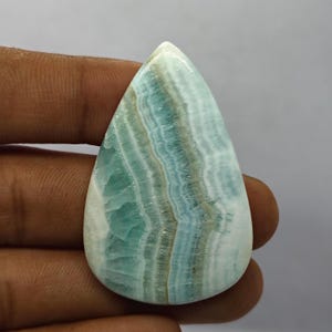 May include: A polished, teardrop-shaped gemstone with a layered pattern of blue, green, and white hues. The stone's surface is smooth and reflective, showcasing natural mineral formations. The gemstone is held by a hand.