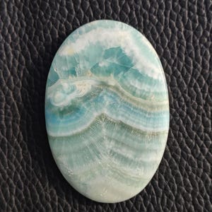 May include: Oval-shaped gemstone with a swirling pattern of blue, white, and pale green hues. The polished surface of the stone displays a natural, layered design, reminiscent of ocean waves. The gemstone is set against a dark, textured background.