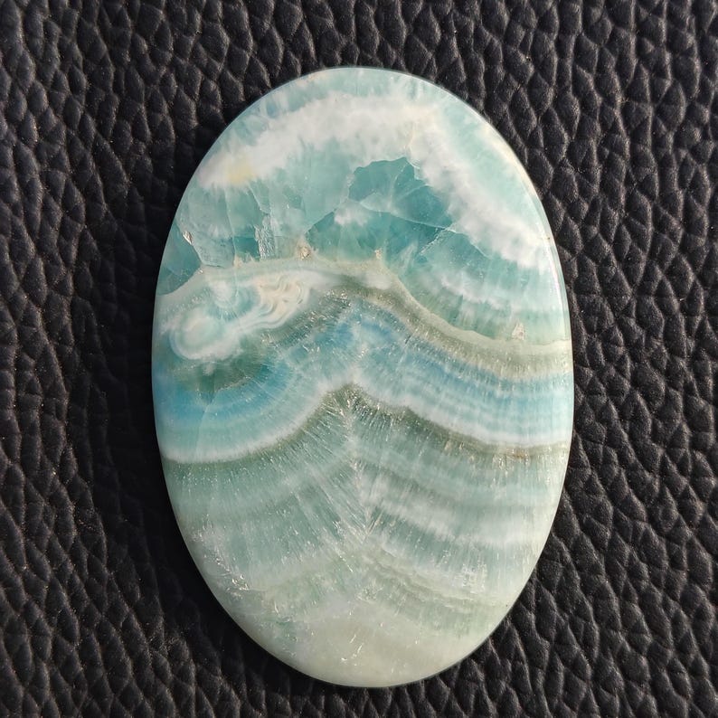 May include: Oval-shaped gemstone with a swirling pattern of blue, white, and pale green hues. The polished surface of the stone displays a natural, layered design, reminiscent of ocean waves. The gemstone is set against a dark, textured background.