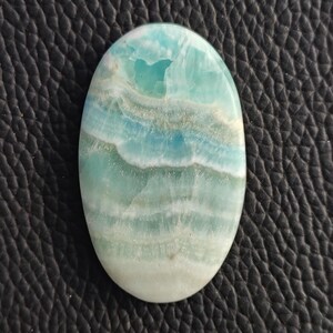 May include: An oval-shaped gemstone with a blend of blue, turquoise, and white hues. The stone has a layered appearance, with bands of color creating a wave-like pattern. The polished surface reflects light, highlighting the natural variations.