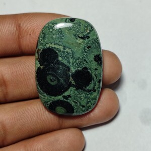 Puede incluir: Gema ovalada con base verde y llamativos patrones circulares negros. La superficie pulida refleja la luz, resaltando el diseño natural único. Adecuada para la creación de joyas, esta piedra mide aproximadamente 3 cm de largo.