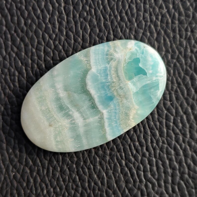 May include: An oval-shaped gemstone with a blend of light blue, white, and teal hues. The stone has a polished surface with a layered, wave-like pattern. The gemstone is set against a dark, textured background.