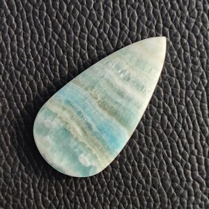 May include: A teardrop-shaped gemstone with a polished surface. The stone features bands of light blue, white, and gray hues. The gemstone is set against a black, textured background, highlighting its natural colors and patterns.