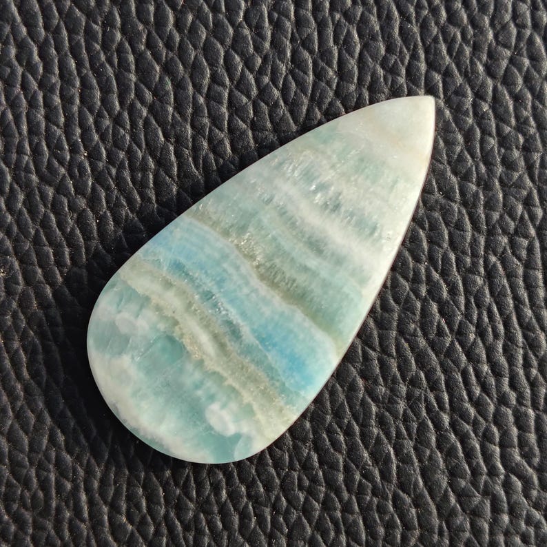 May include: A teardrop-shaped gemstone with a polished surface. The stone features bands of light blue, white, and gray hues. The gemstone is set against a black, textured background, highlighting its natural colors and patterns.