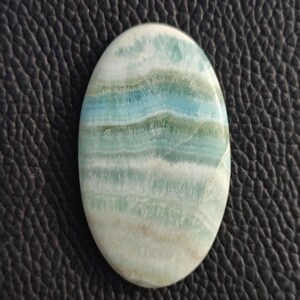 May include: Oval-shaped gemstone with a layered pattern of white, light blue, and seafoam green hues. The polished stone has a smooth surface and a natural, organic appearance. The gemstone is set against a dark, textured background.