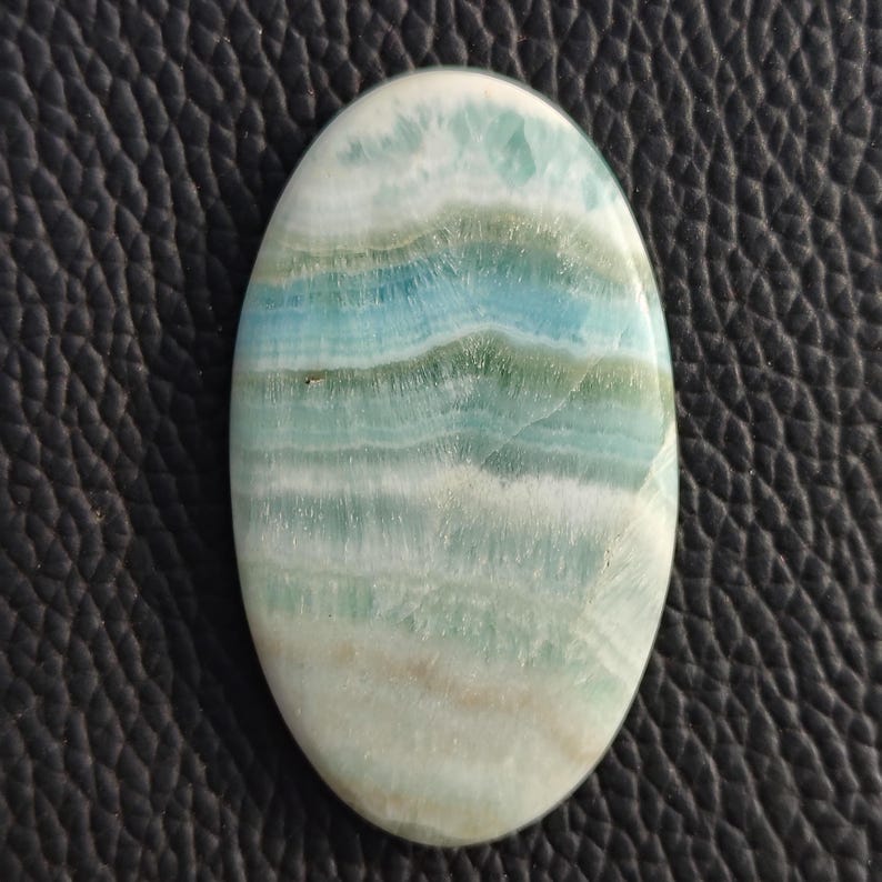 May include: Oval-shaped gemstone with a layered pattern of white, light blue, and seafoam green hues. The polished stone has a smooth surface and a natural, organic appearance. The gemstone is set against a dark, textured background.