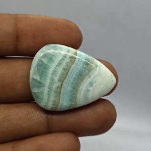 Pode incluir: Uma pedra preciosa polida em forma de gota, com uma mistura de faixas azul p&aacute;lido, brancas e castanhas. A pedra tem uma superf&iacute;cie lisa e brilhante e um aspeto natural e org&acirc;nico. A pedra &eacute; segurada numa m&atilde;o.