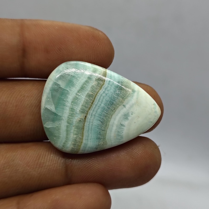 Pode incluir: Uma pedra preciosa polida em forma de gota, com uma mistura de faixas azul p&aacute;lido, brancas e castanhas. A pedra tem uma superf&iacute;cie lisa e brilhante e um aspeto natural e org&acirc;nico. A pedra &eacute; segurada numa m&atilde;o.