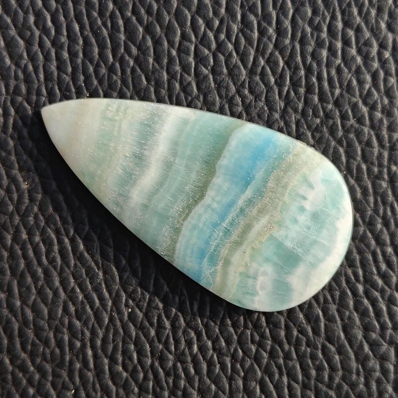 May include: A polished, teardrop-shaped gemstone with a swirling pattern of light blue, turquoise, and white. The stone has a smooth, glossy surface and is set against a textured black background. The gemstone is likely used for jewelry or decorative purposes.