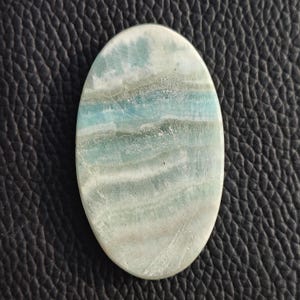 May include: Oval-shaped gemstone with a polished surface. The stone features a blend of light blue, white, and gray hues, creating a layered, wave-like pattern. The gemstone is set against a dark, textured background.