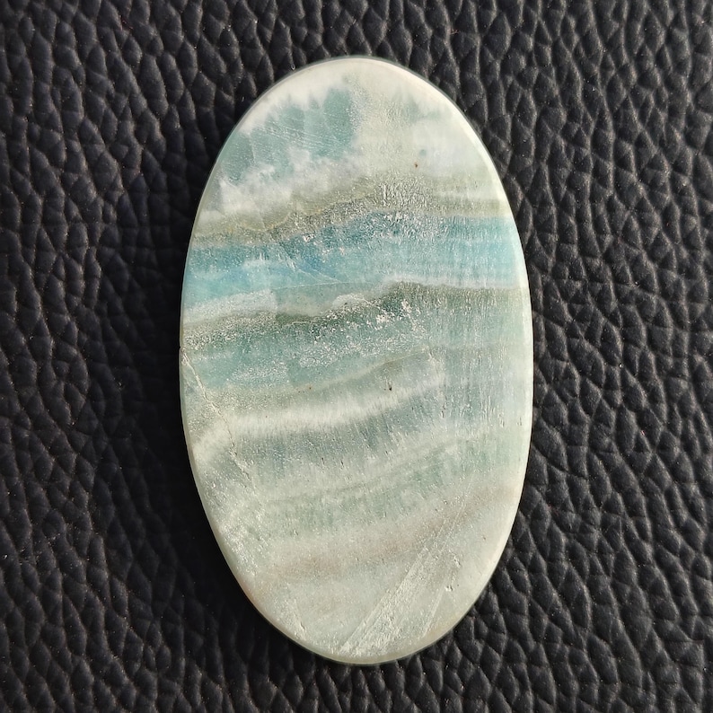 May include: Oval-shaped gemstone with a polished surface. The stone features a blend of light blue, white, and gray hues, creating a layered, wave-like pattern. The gemstone is set against a dark, textured background.