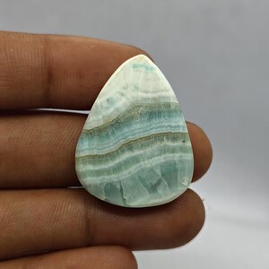 Pode incluir: Uma gema em forma de gota com um padr&atilde;o em camadas de faixas azul claro, brancas e castanhas. A pedra polida tem uma superf&iacute;cie lisa e uma apar&ecirc;ncia ligeiramente transl&uacute;cida.