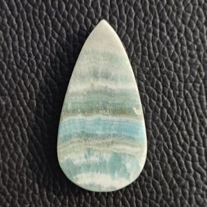 May include: A teardrop-shaped gemstone with a polished surface. The stone features bands of light blue, green, and white, creating a layered effect. The gemstone is set against a textured black background.