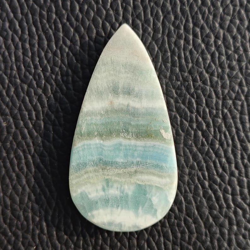 May include: A teardrop-shaped gemstone with a polished surface. The stone features bands of light blue, green, and white, creating a layered effect. The gemstone is set against a textured black background.