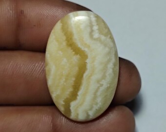 Puede incluir: Gema ovalada con superficie pulida. La piedra presenta un color base blanco cremoso con bandas fluidas de tonos amarillo claro y marrón claro. La gema se sostiene en una mano, mostrando su tamaño y forma.