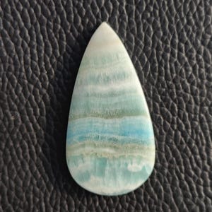 May include: A teardrop-shaped gemstone with a polished surface. The stone features layers of white, light green, and blue hues. The gemstone is set against a dark, textured background.