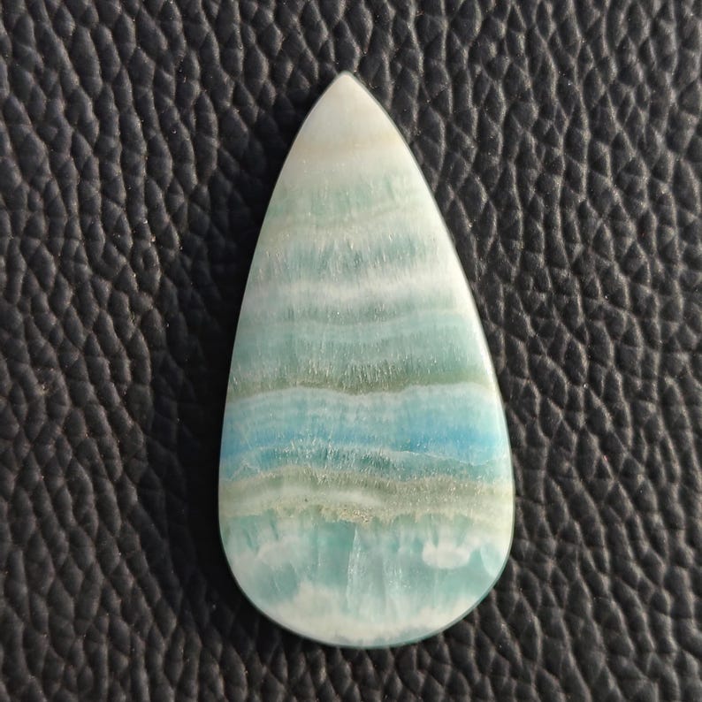 May include: A teardrop-shaped gemstone with a polished surface. The stone features layers of white, light green, and blue hues. The gemstone is set against a dark, textured background.
