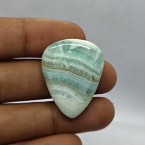 Pode incluir: Uma pedra preciosa polida em forma de gota, com um padr&atilde;o em camadas de tons azul p&aacute;lido, branco e bege. A pedra &eacute; segurada na m&atilde;o, mostrando sua beleza natural e varia&ccedil;&otilde;es de cores &uacute;nicas. A pedra preciosa &eacute; provavelmente usada para joias.
