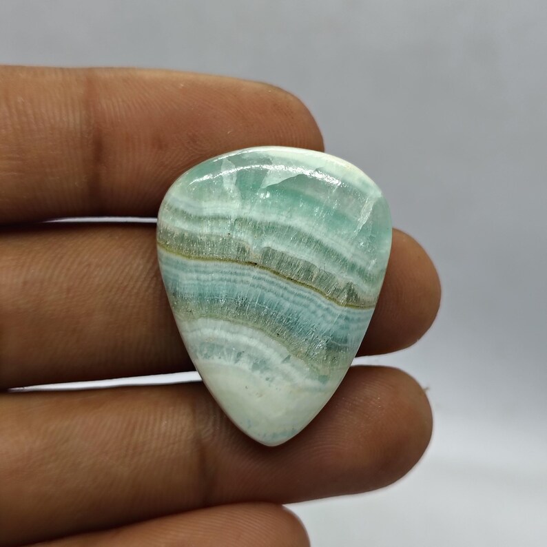 Pode incluir: Uma pedra preciosa polida em forma de gota, com um padr&atilde;o em camadas de tons azul p&aacute;lido, branco e bege. A pedra &eacute; segurada na m&atilde;o, mostrando sua beleza natural e varia&ccedil;&otilde;es de cores &uacute;nicas. A pedra preciosa &eacute; provavelmente usada para joias.
