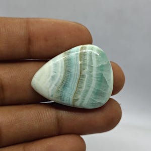 Pode incluir: Uma pedra preciosa polida em forma de gota, com uma mistura de faixas brancas, azul claro e castanhas. A pedra tem uma superf&iacute;cie lisa e brilhante e &eacute; segurada na m&atilde;o contra um fundo neutro. &Eacute; provavelmente um caboch&atilde;o.