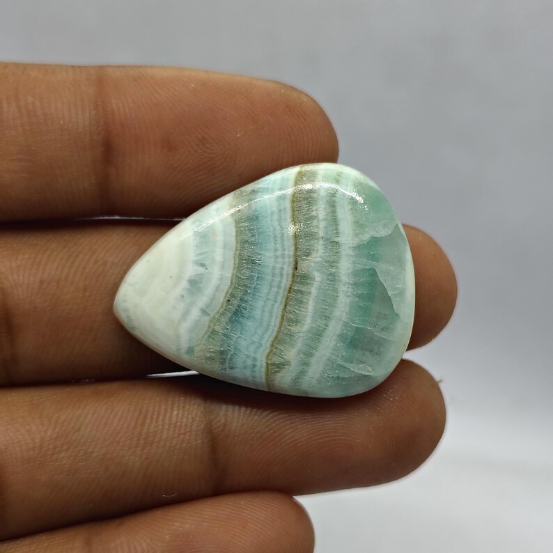 Pode incluir: Uma pedra preciosa polida em forma de gota, com uma mistura de faixas brancas, azul claro e castanhas. A pedra tem uma superf&iacute;cie lisa e brilhante e &eacute; segurada na m&atilde;o contra um fundo neutro. &Eacute; provavelmente um caboch&atilde;o.