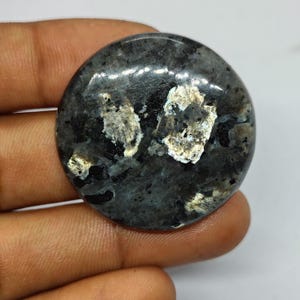 May include: A circular, polished gemstone with a dark grey base and shimmering silver and gold inclusions. The stone has a smooth, glossy finish. The gemstone is likely a cabochon, suitable for jewellery making. It is held in a hand.