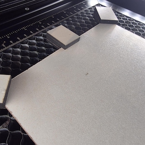 Könnte beinhalten: Ein hellgraues rechteckiges Material befindet sich in einer Laserschneidmaschine. Drei rechteckige Blöcke sind auf dem Material platziert, wahrscheinlich um es während des Schneidens zu halten. Die Maschine hat ein schwarzes Wabenmuster.