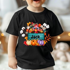 Könnte beinhalten: Schwarzes T-Shirt mit einem Thanksgiving-Truthahn-Design, Kürbissen, Blättern und dem Namen "Jack" auf einem blauen Holzschild. Der Truthahn hat ein Patchwork-Design und ist von Baumwolle und Herbstlaub umgeben.