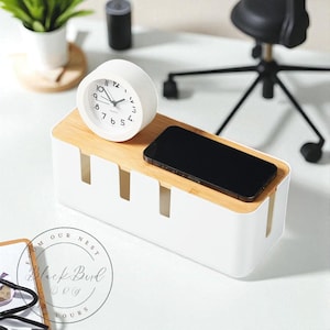 Puede incluir: Un organizador de cables blanco con una tapa de bambú, que contiene un teléfono inteligente negro y un reloj analógico blanco. El reloj marca las 9:10. El organizador tiene ranuras para la gestión de cables.